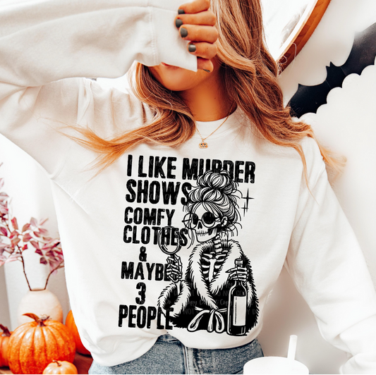 White I Like Murder Shows sweatshirt with black skeleton in robe holding wine and magnifying glass, worn by a woman in a cozy home setting.