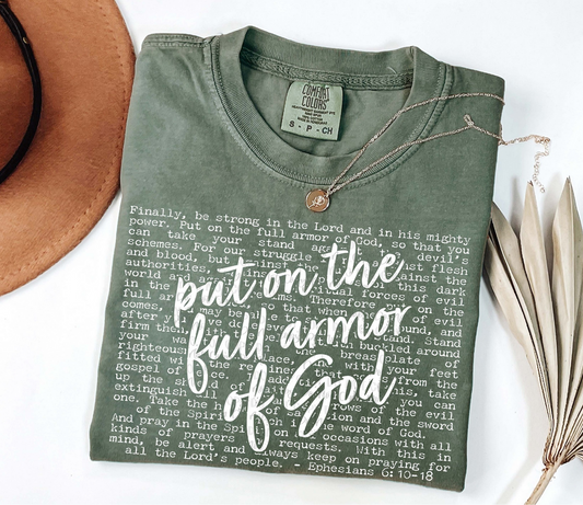 Olive green Put on the Full Armor of God t-shirt with Ephesians 6:10–18 scripture text and bold overlay lettering, laid flat with jewelry and hat.