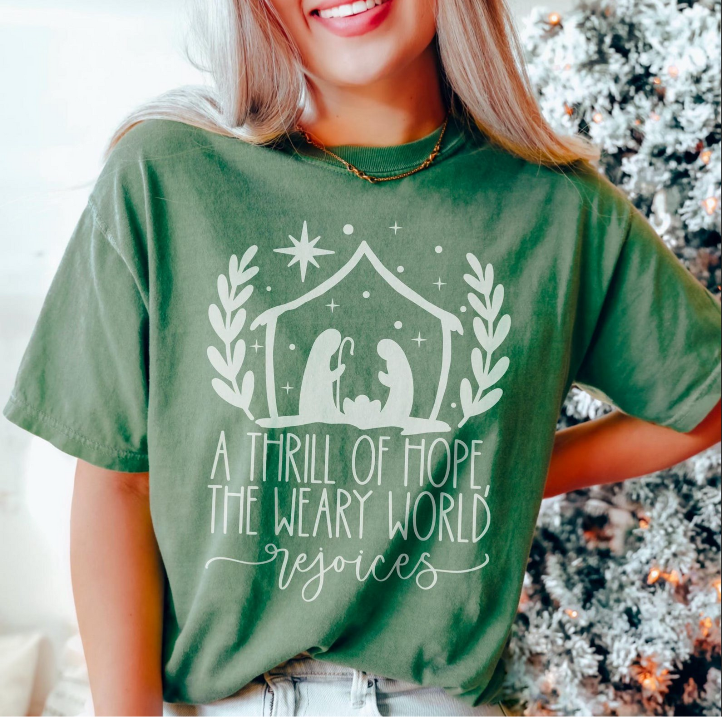 Green t-shirt with nativity design and text “A Thrill of Hope, The Weary World Rejoices” in white lettering, modeled by woman standing near Christmas tree