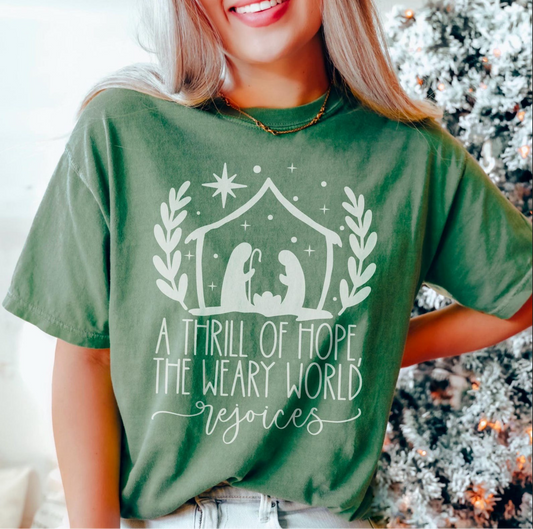 Green t-shirt with nativity design and text “A Thrill of Hope, The Weary World Rejoices” in white lettering, modeled by woman standing near Christmas tree