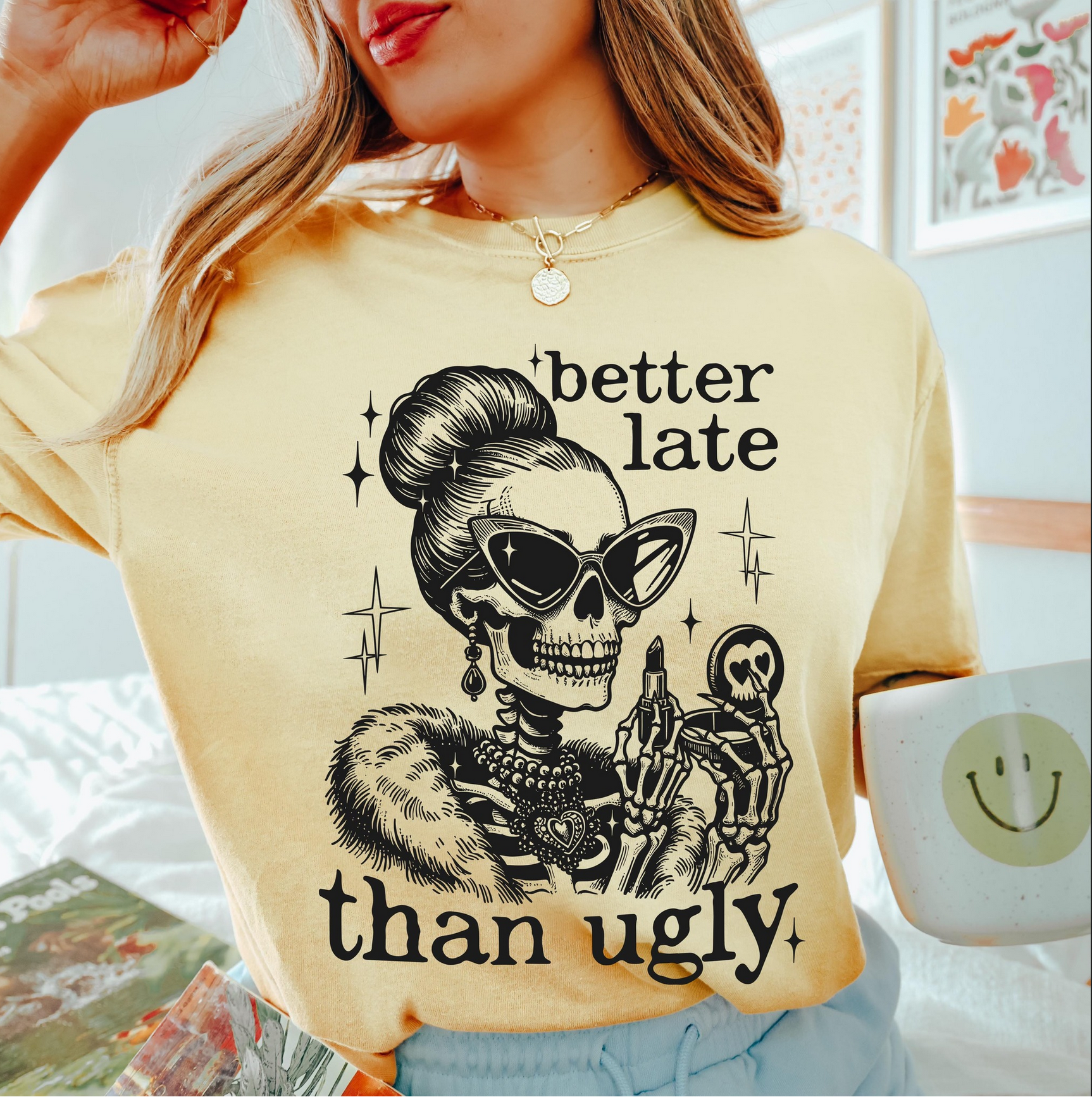 Light yellow Better Late Than Ugly t-shirt with stylish skeleton in sunglasses and fur coat holding a compact mirror, worn by a woman holding a smiley mug.