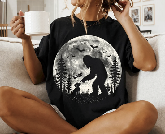 Model wearing a black graphic T-shirt featuring Bigfoot booping a dog beneath a full moon with pine trees and flying bats.