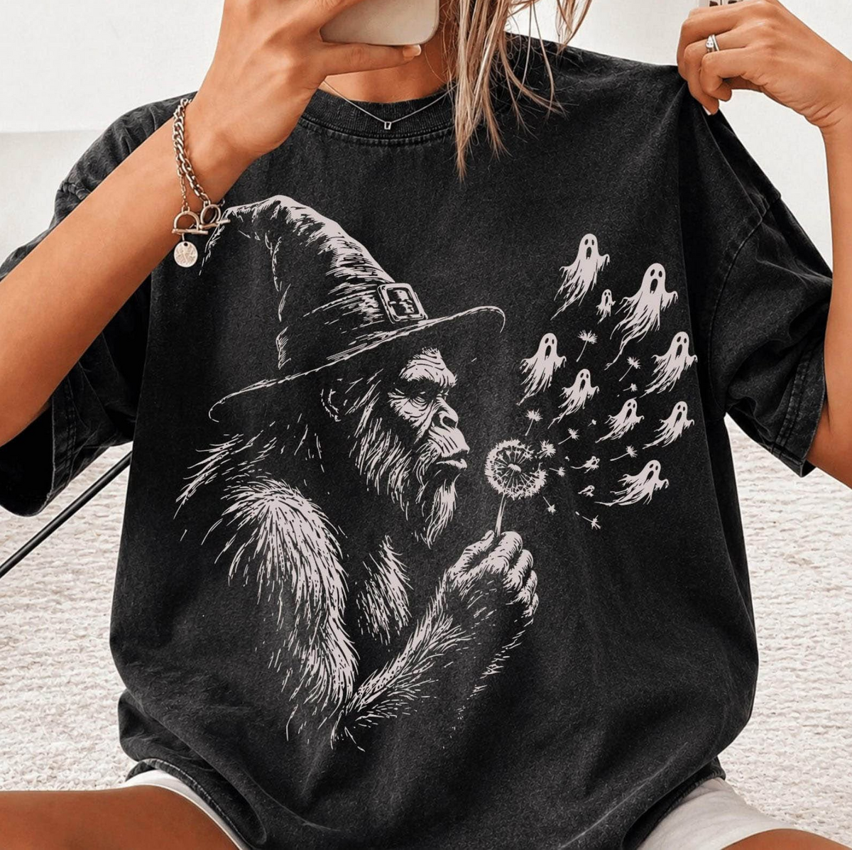 Model wearing a black graphic T-shirt featuring Bigfoot in a witch hat blowing a dandelion that transforms into small flying ghosts.