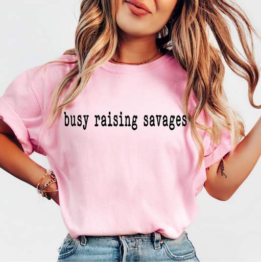 Pink Busy Raising Savages t-shirt with black text, worn by a woman with long wavy hair and casual denim shorts.