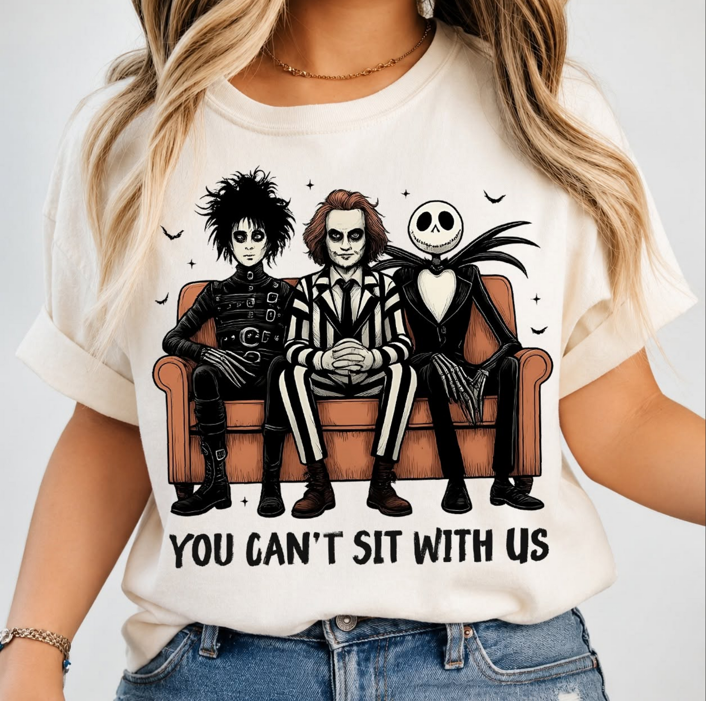Cream graphic t-shirt featuring a spooky gothic trio sitting on a couch with the phrase "You Can't Sit With Us."