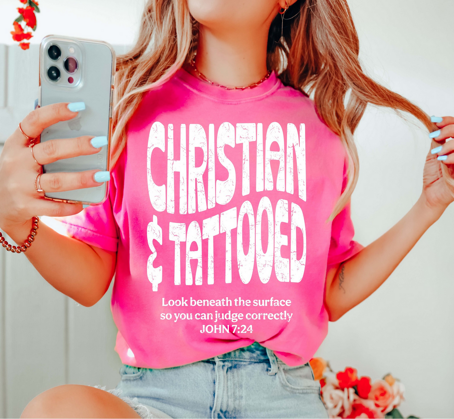 Bright pink Christian & Tattooed t-shirt with distressed white lettering and John 7:24 Bible verse, worn by a woman holding her phone.