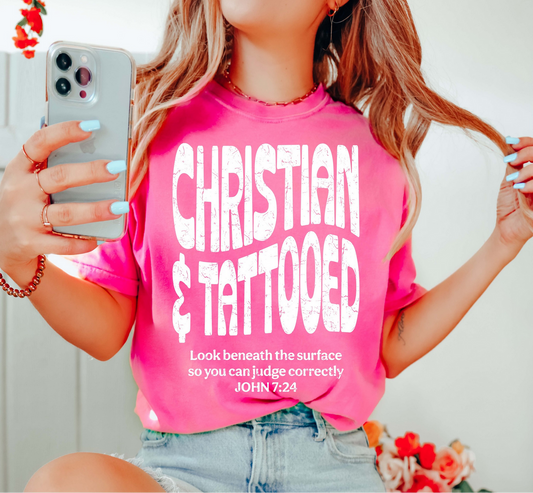 Bright pink Christian & Tattooed t-shirt with distressed white lettering and John 7:24 Bible verse, worn by a woman holding her phone.