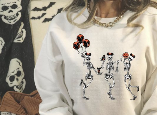 White sweatshirt with three dancing skeletons — one holding orange balloons, one with a witch hat, and one with a pumpkin head. Halloween design.