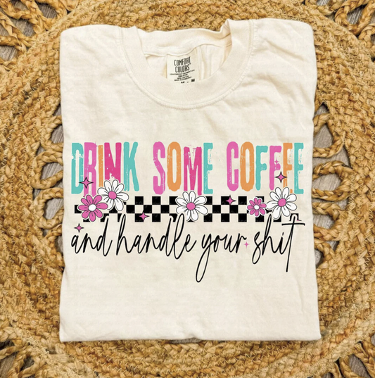 Cream Drink Some Coffee and Handle Your Sh*t t-shirt with colorful retro lettering, floral details, and checkered design, laid flat on a woven mat.
