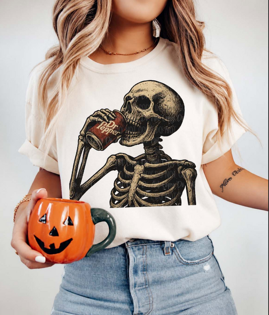 Vintage skeleton drinking a Dr Pepper while holding a pumpkin mug on a cream graphic t-shirt for Halloween and fall.