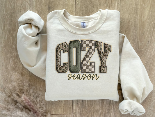 Folded cream sweatshirt with the phrase Cozy Season in neutral patchwork lettering.