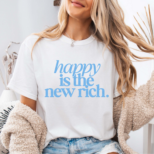 White Happy Is the New Rich t-shirt with blue retro lettering, worn by a woman with long hair and cozy cardigan, holding a coffee mug.
