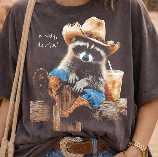 Vintage western graphic t-shirt featuring a raccoon wearing a cowboy hat and boots with the phrase Howdy Darlin and iced coffee beside it