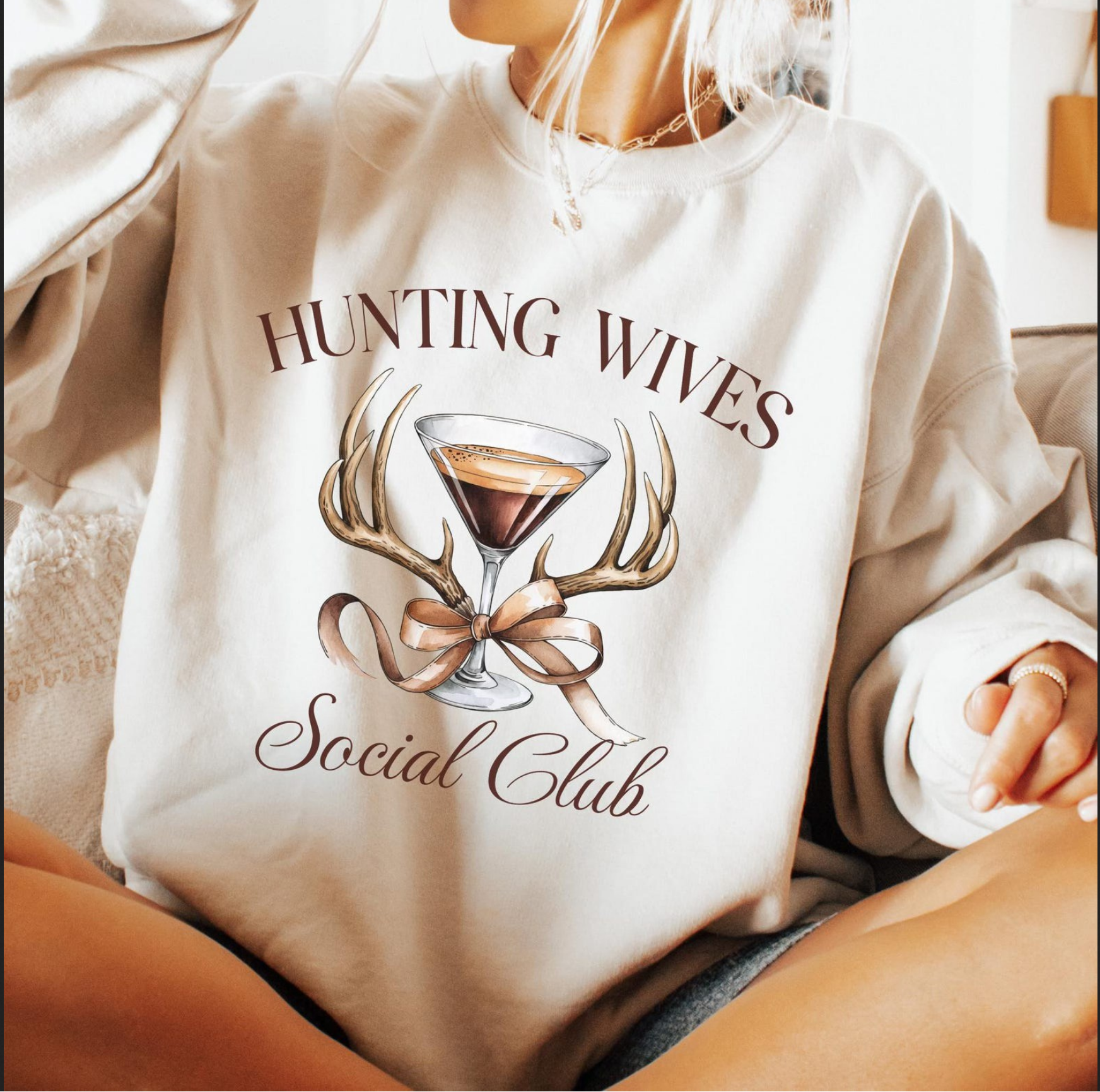Beige sweatshirt with “Hunting Wives Social Club” text and martini glass framed by antlers and ribbon