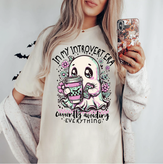 Cream In My Introvert Era t-shirt with cartoon ghost holding coffee, flowers, and pastel accents, worn by a woman with a cozy cardigan.