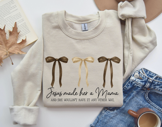 Cream Jesus Made Her a Mama sweatshirt with three neutral-tone bows and faith-based text, laid flat next to jeans, coffee, and fall decor.