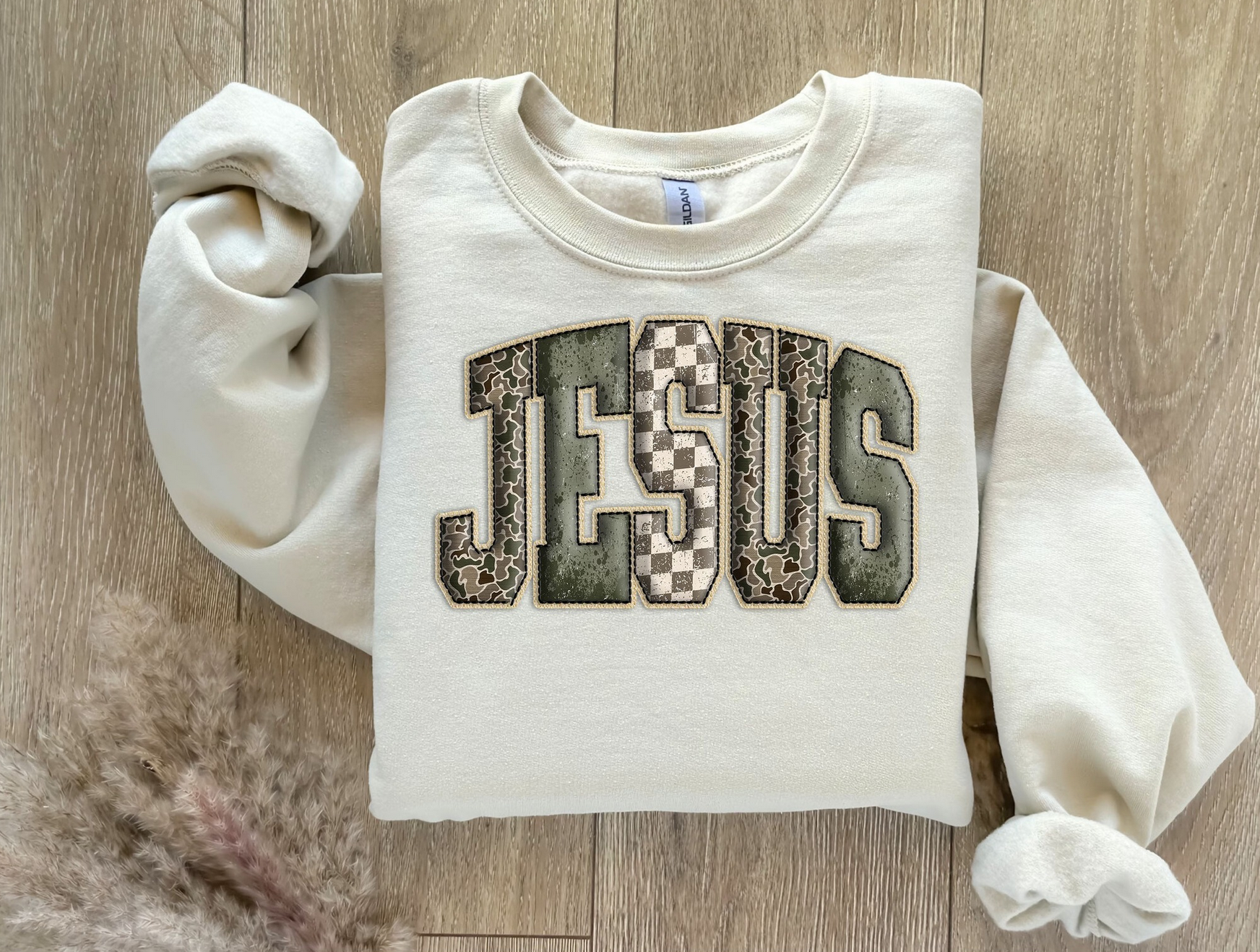 Folded cream sweatshirt with large JESUS patchwork letters in checkered and textured patterns.