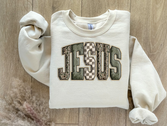 Folded cream sweatshirt with large JESUS patchwork letters in checkered and textured patterns.