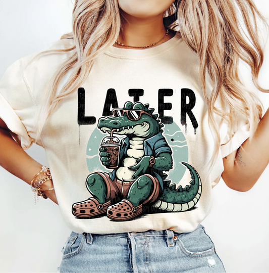 Cream Later Alligator t-shirt with cartoon gator in sunglasses sipping a drink, wearing crocs and jacket, worn casually with denim shorts.
