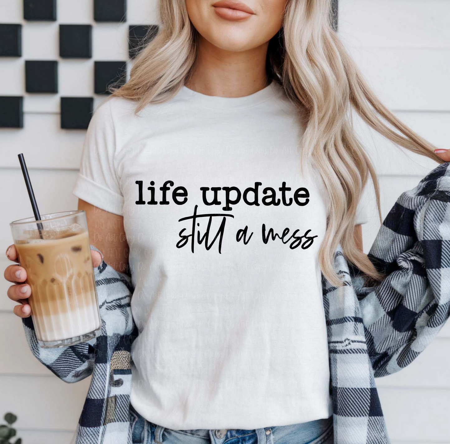 White Life Update Still a Mess t-shirt with black text, worn by a woman holding iced coffee and wearing a plaid shirt over it.