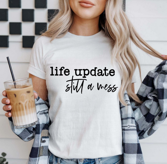 White Life Update Still a Mess t-shirt with black text, worn by a woman holding iced coffee and wearing a plaid shirt over it.