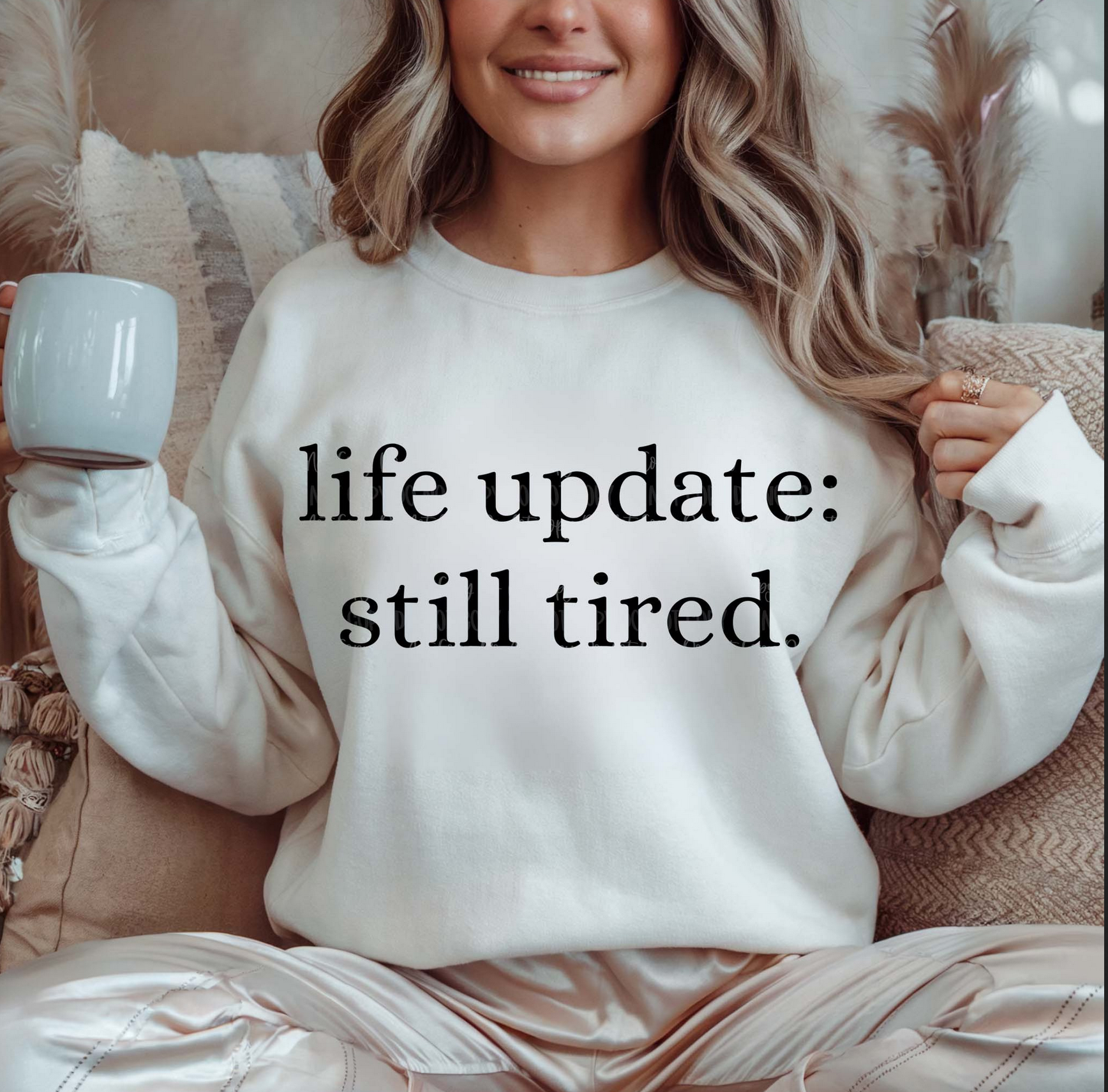 Cream Life Update Still Tired sweatshirt with black text, worn by a woman sitting indoors holding a coffee mug.