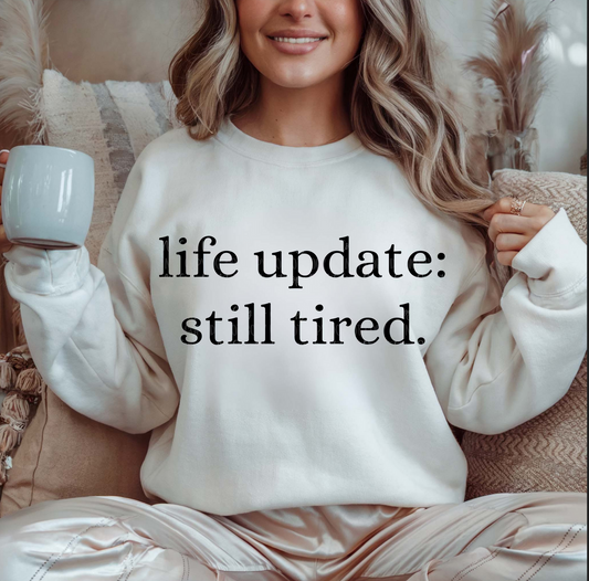 Cream Life Update Still Tired sweatshirt with black text, worn by a woman sitting indoors holding a coffee mug.