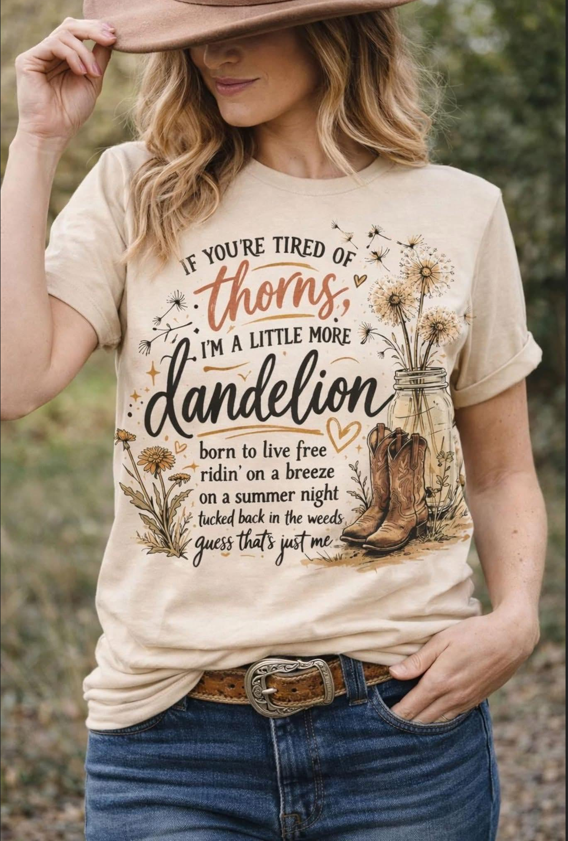Model wearing a beige western graphic T-shirt featuring dandelions, cowboy boots, a mason jar, and the saying If You’re Tired of Thorns, I’m a Little More Dandelion.