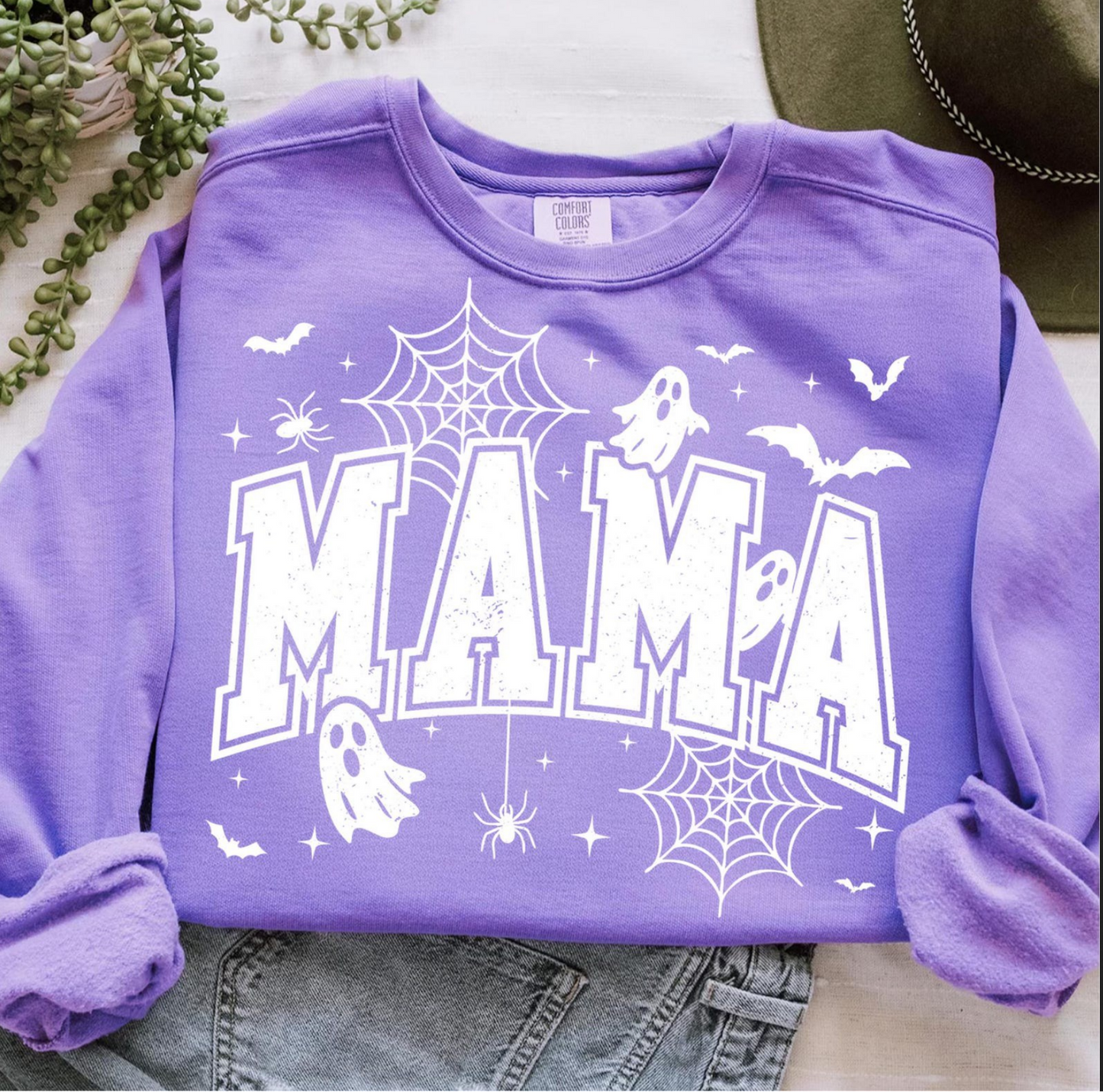 Purple crewneck sweatshirt with large white “MAMA” varsity lettering decorated with ghosts, spider webs, bats, and stars.