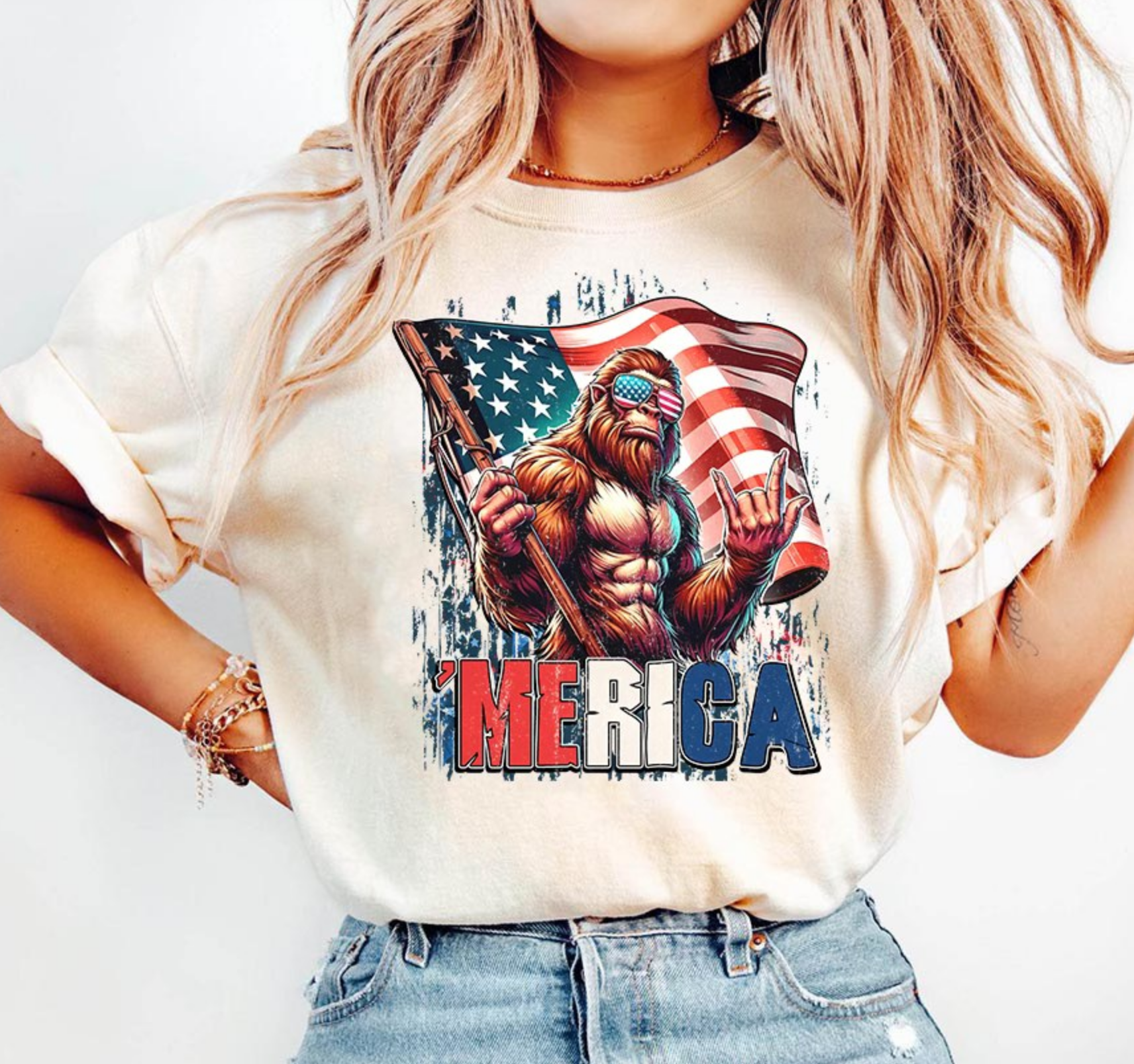 Beige t-shirt with Bigfoot wearing American flag sunglasses holding flag with text ‘Merica