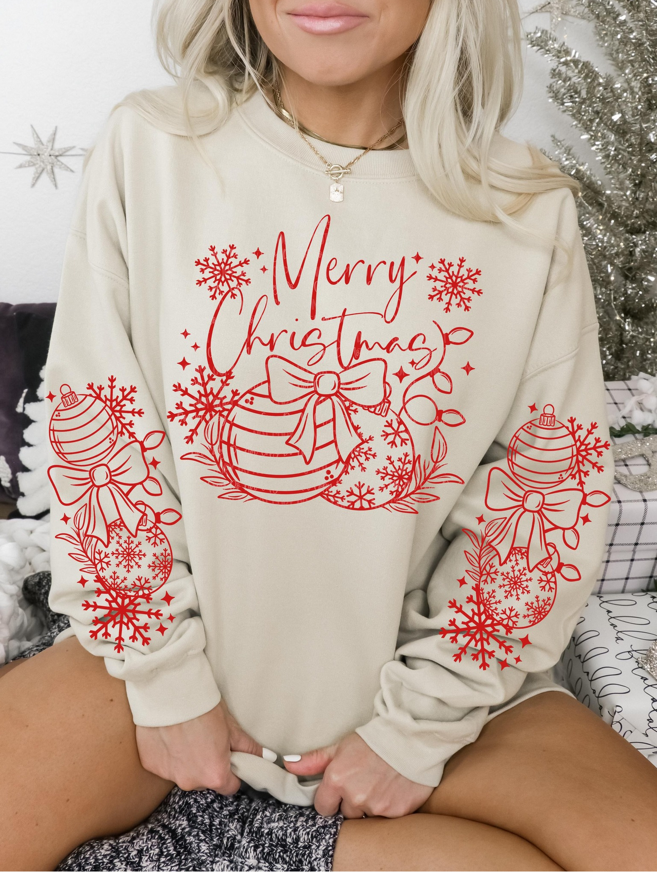 Beige sweatshirt with red “Merry Christmas” lettering, bows, ornaments, and snowflake sleeve details, modeled by woman sitting by Christmas décor