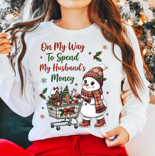 Model wearing a white Christmas sweatshirt featuring a snowwoman pushing a gift-filled shopping cart with the saying On My Way to Spend My Husband’s Money.