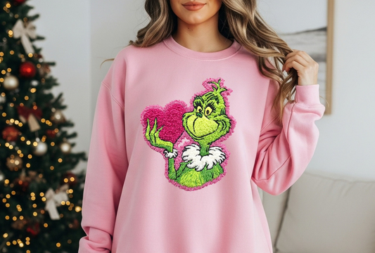 Pink sweatshirt with a fuzzy green holiday character holding a bright pink heart.