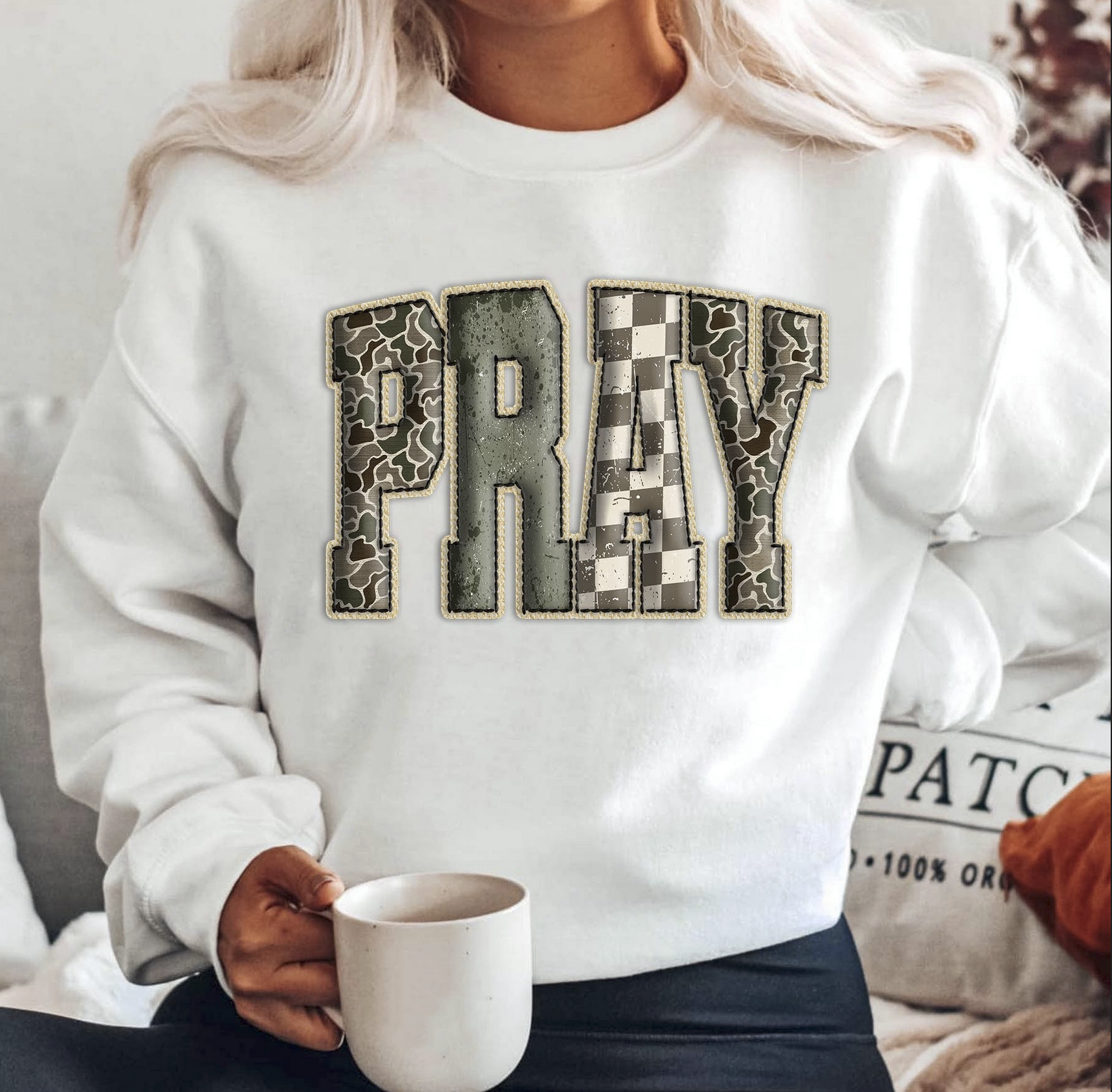 White sweatshirt with large patchwork PRAY lettering featuring camo and checkered patterns.
