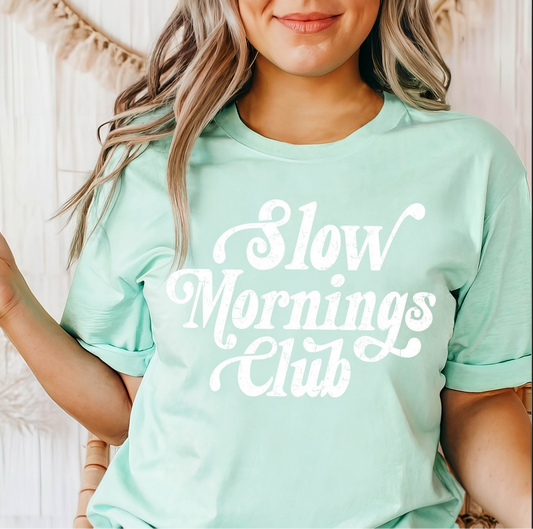 Mint green Slow Mornings Club t-shirt with white retro lettering, worn by a woman with loose waves, sitting casually indoors.
