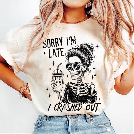 Cream Sorry I’m Late I Crashed Out t-shirt with black skeleton holding coffee graphic, worn by a woman with wavy hair and rolled sleeves.