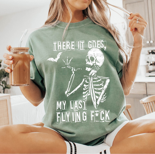 Sage green There It Goes My Last Flying F*ck t-shirt with white skeleton and bat graphic, worn by a woman holding iced coffee in a casual home setting.