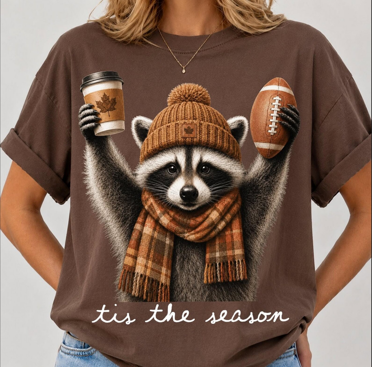 Model wearing a brown graphic T-shirt featuring a raccoon in a knit hat and plaid scarf holding a football and coffee beneath the words ’Tis the Season.