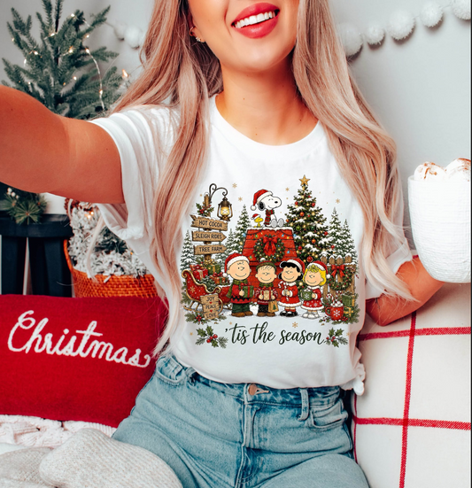 Model wearing a white Christmas graphic T-shirt featuring Snoopy, Woodstock, Charlie Brown, and Peanuts friends gathered around a decorated doghouse and tree.