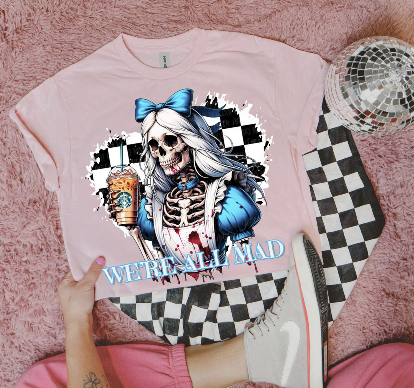 Pink tee with skeleton girl in blue bow holding iced coffee, text “We’re All Mad.”
Close-up of spooky gothic parody skeleton graphic on pink shirt.