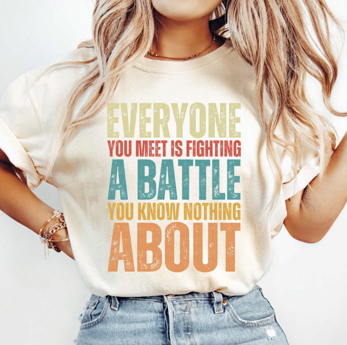Cream Everyone Is Fighting a Battle t-shirt with retro multi-color bold lettering, worn by a woman with casual denim shorts.